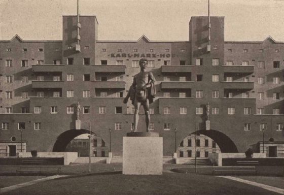 The long symmetrical façade of the Karl-Marx-Hof, its name in raised lettering on the wall between two tall flag towers, with a bronze statue of a standing nude man on a stone plinth in the courtyard in front of it.