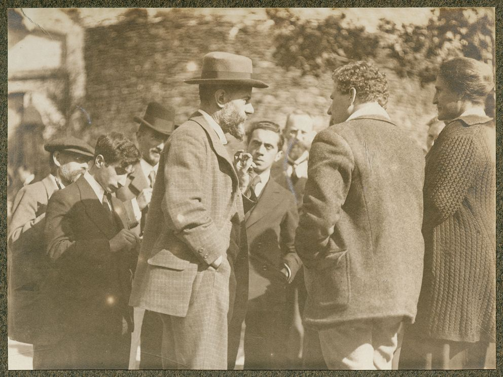 A group of men in conversation outdoors in front of an ivy-covered wall; Max Weber, bearded and in a broad-brimmed hat and a checked suit, stands in profile at the centre, speaking, while a young man beside him faces the camera