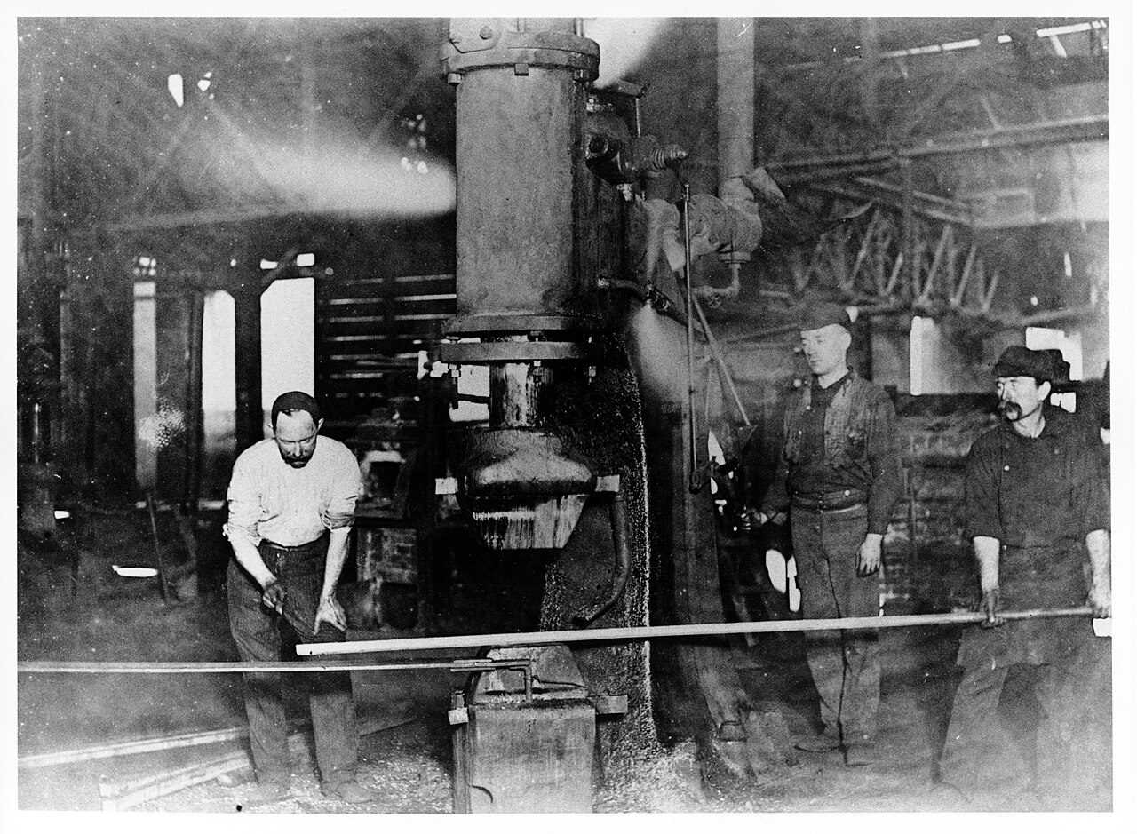 Three workmen in a dim forge shop stand around a large steam hammer; two of them hold a long steel bar on the anvil while the third reaches for a tool