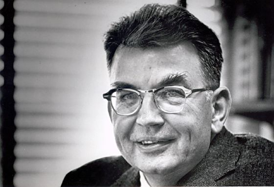 Ronald Coase in middle age, in glasses and a tweed jacket, smiling slightly, photographed indoors in front of a window blind and a bookshelf