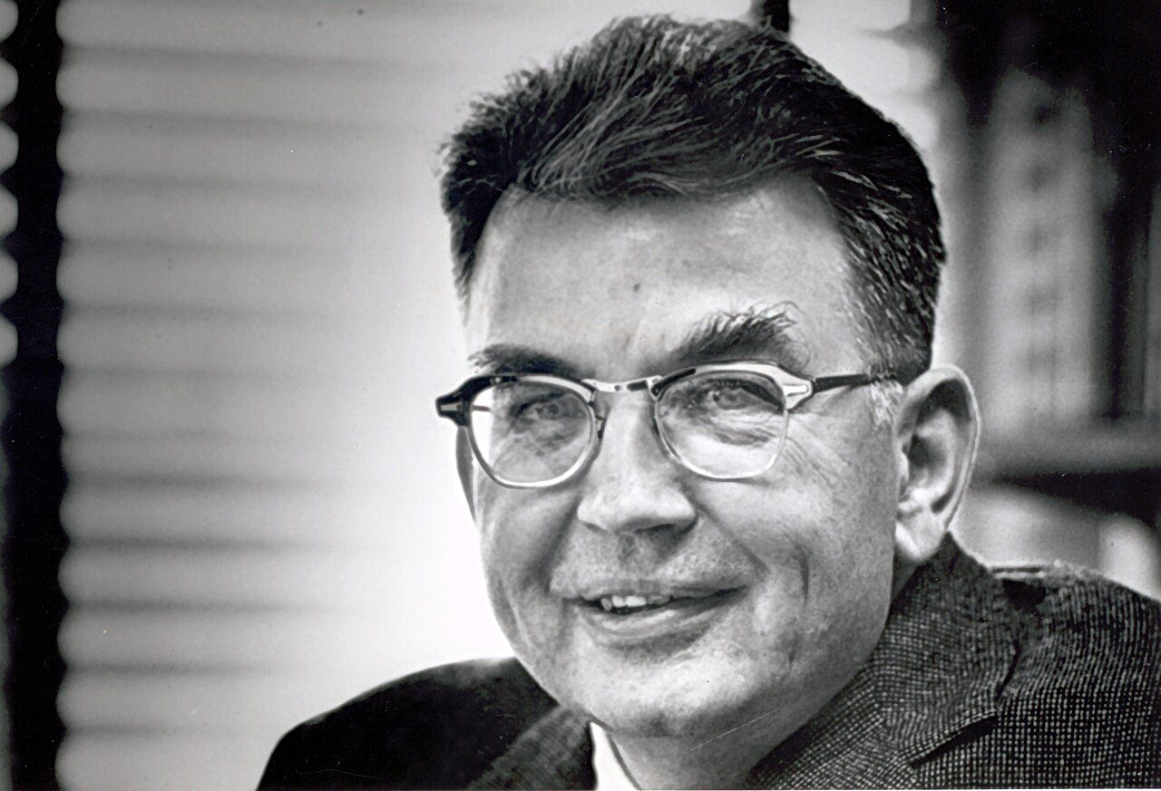 Ronald Coase in middle age, in glasses and a tweed jacket, smiling slightly, photographed indoors in front of a window blind and a bookshelf