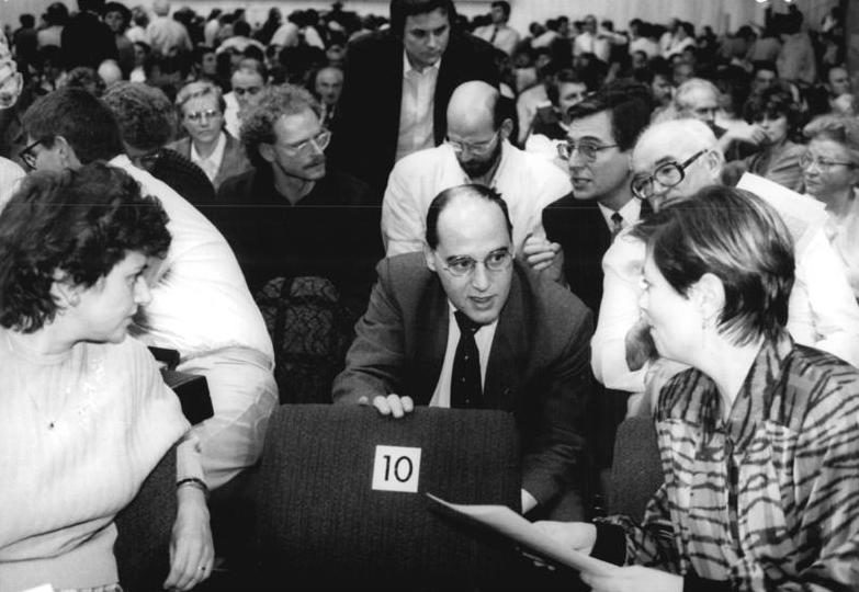 A bespectacled Gregor Gysi in a dark suit leans over the back of a row of seats in a crowded congress hall, talking with delegates pressed around him; the seat in front of him carries a card numbered 10.